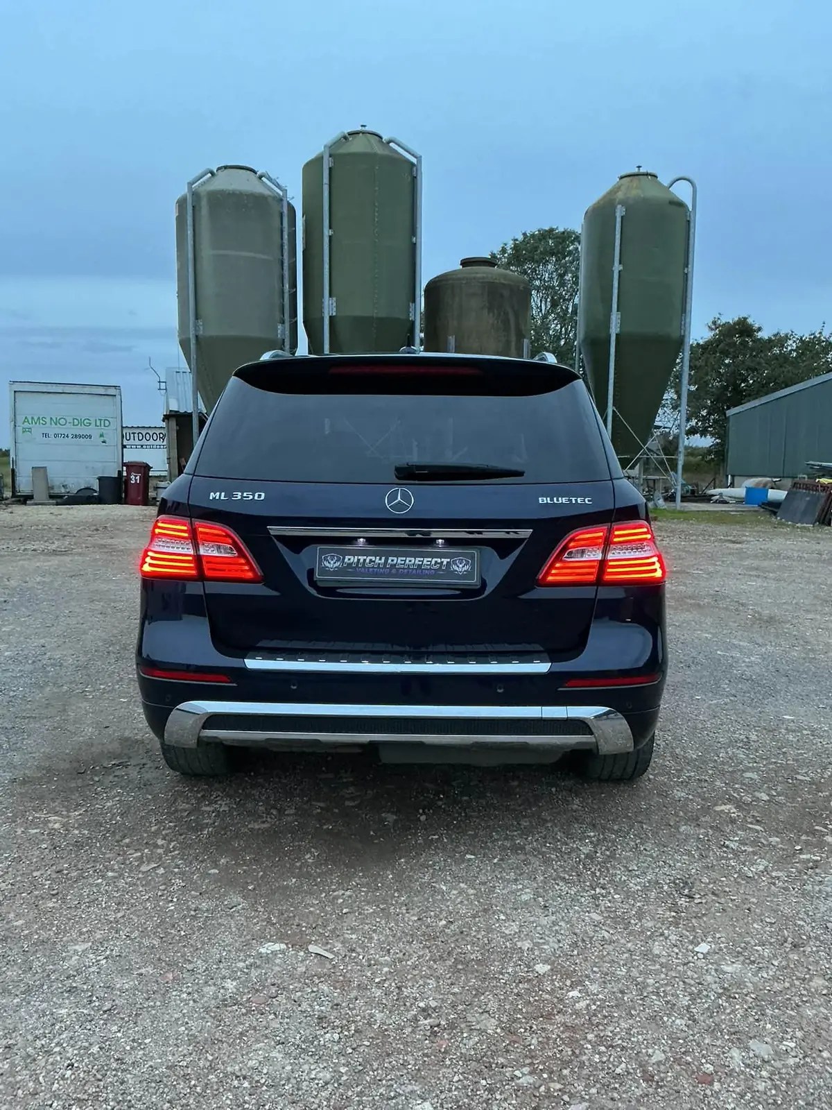 Clean dark Mercedes ML350 rear on gravel in front of farm silos
