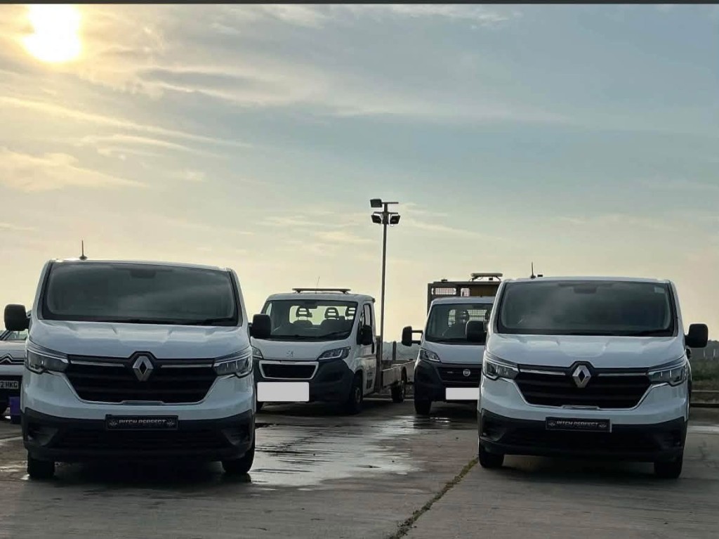 Several white vans with Pitch Perfect plates on a wet forecourt at sunset